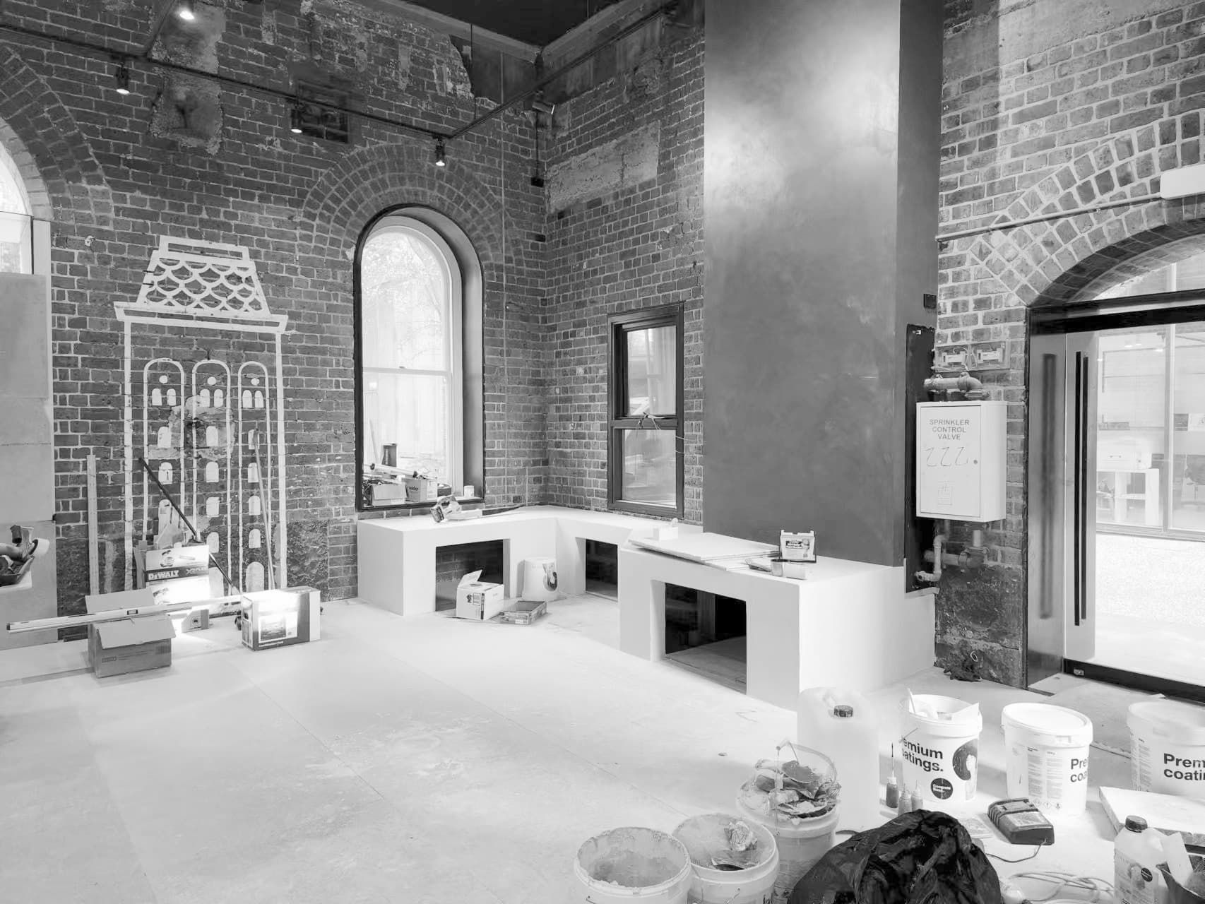 ULA Café in Collingwood during works, with a trowelled microcement column and bench forms against exposed brick walls, in black and white